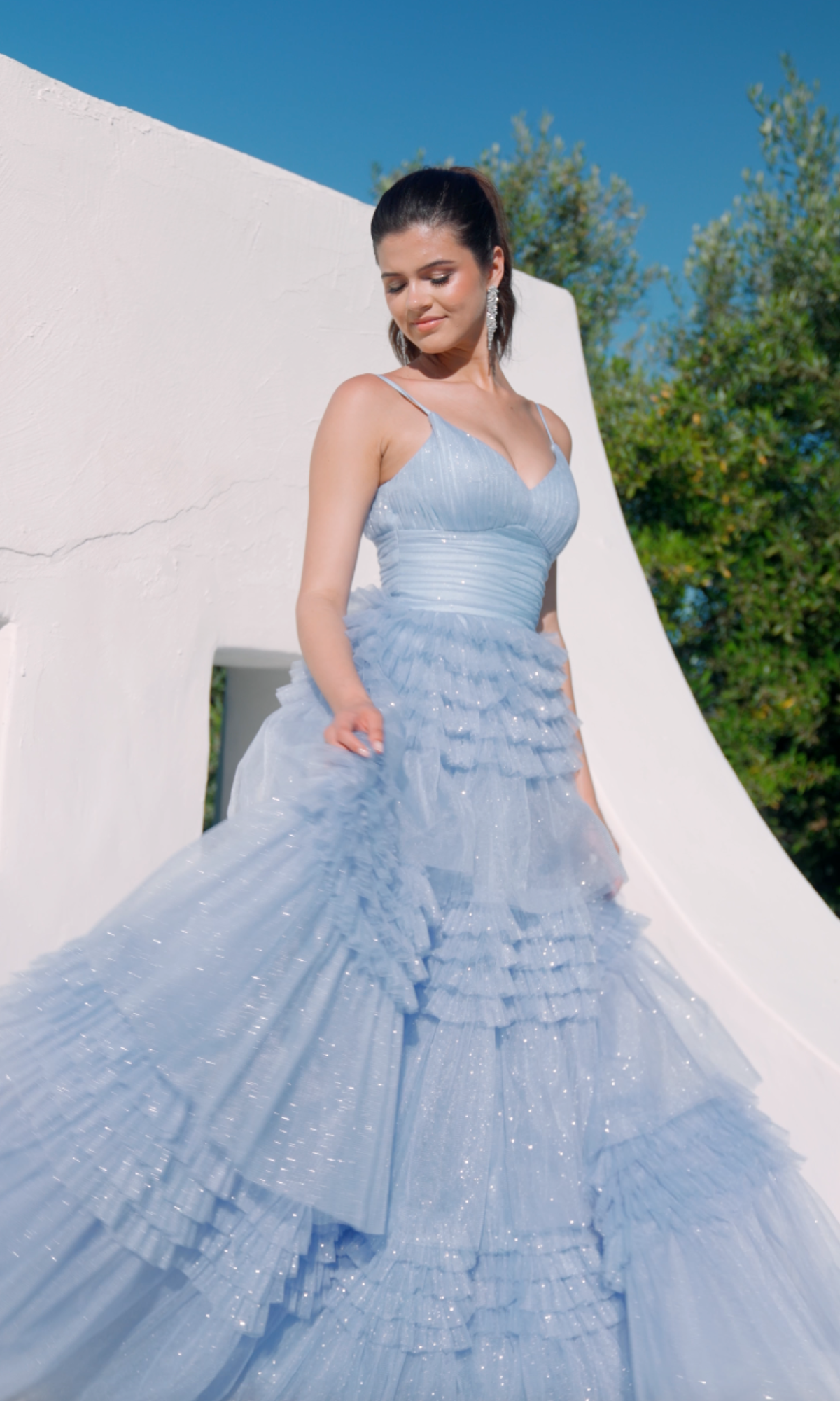A woman wearing the Eureka 9888 Lace-Up Long Glitter-Tulle Prom Dress stands outdoors before a white curved structure, with greenery in the background.